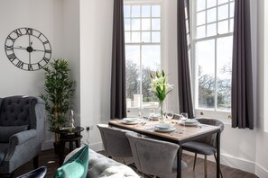 A dining area with a wooden table set for two, a grey sofa, a large wall clock, and a window with curtains.