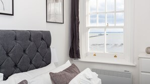 A bedroom with a tufted headboard, a bed with white and brown bedding, a window with a view of the sea, and a white nightstand with a lamp. - The Regency Apartments (Southend-on-Sea)