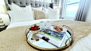 Un lit bien fait, aux couleurs blanches et beige, un plateau avec des articles pour le petit-déjeuner et un magazine posés sur le lit.