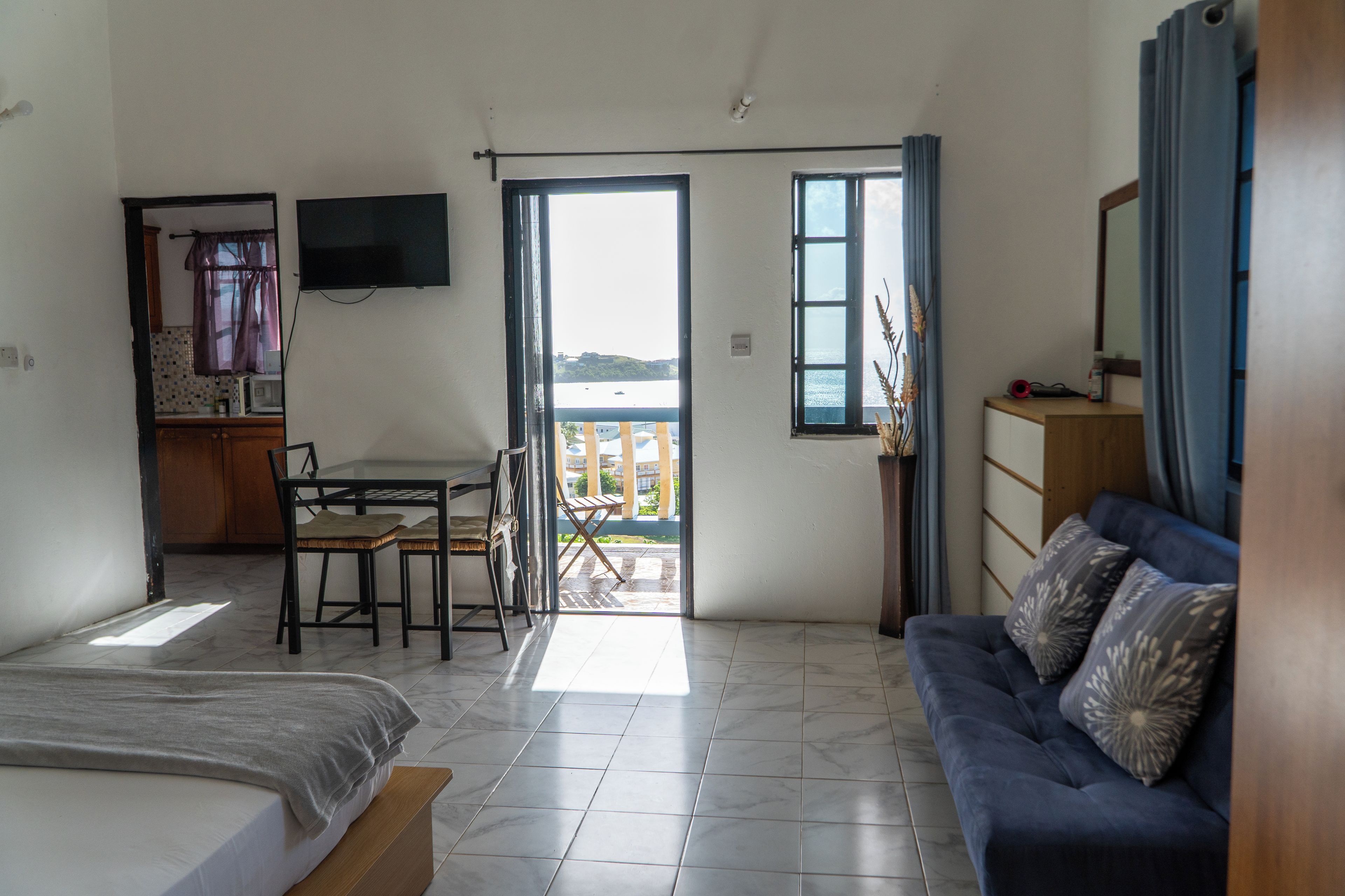 Una habitación con cama, sofá, mesa de comedor con sillas, televisor montado en la pared y balcón con vistas.