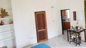 Een kamer met een houten deur, een witte muur, een betegelde vloer, een eettafel met stoelen en een klein plankje met decoratieve objecten.