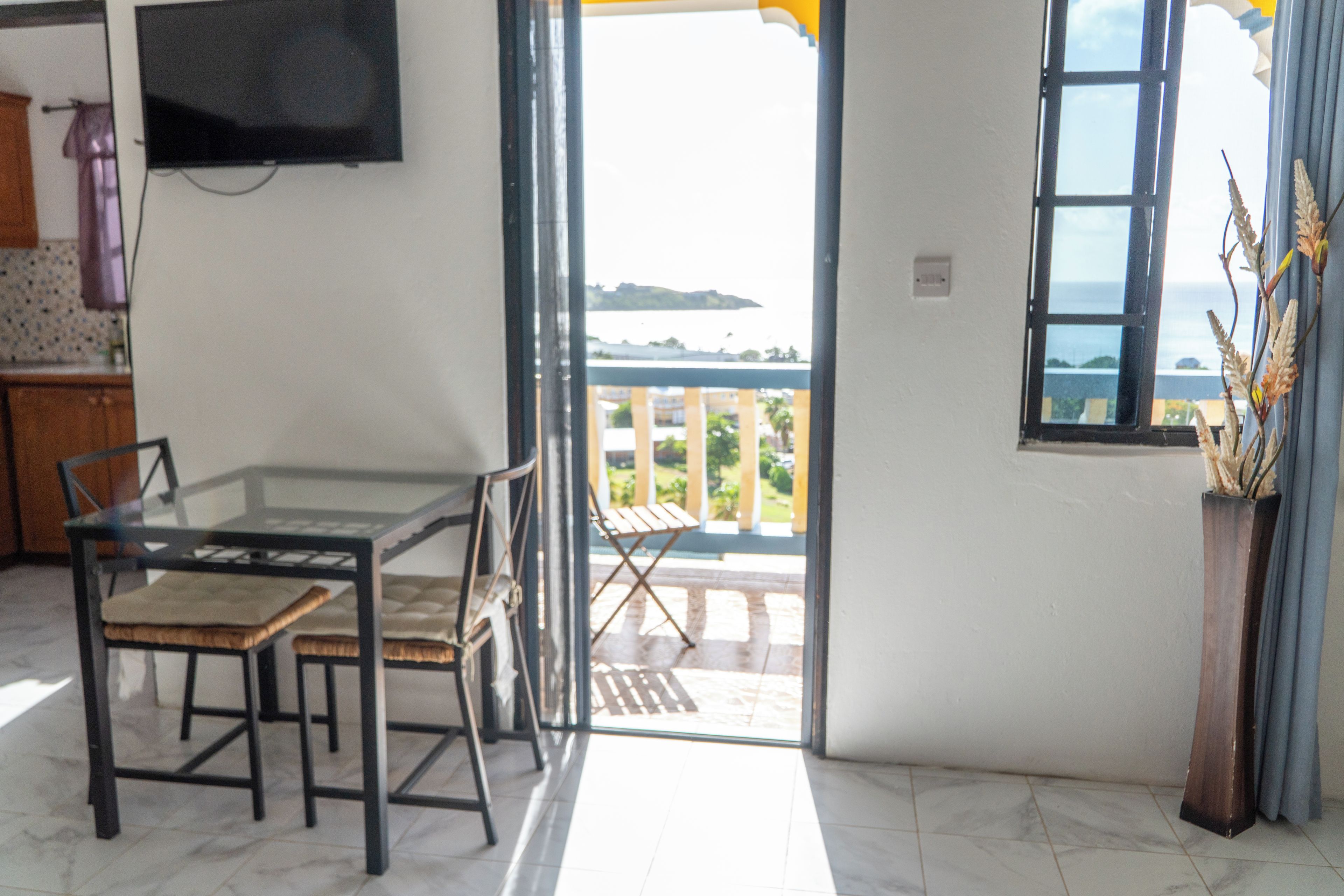 Una habitación con una mesa de comedor y sillas, un televisor montado en la pared y vista al mar a través de una puerta abierta.