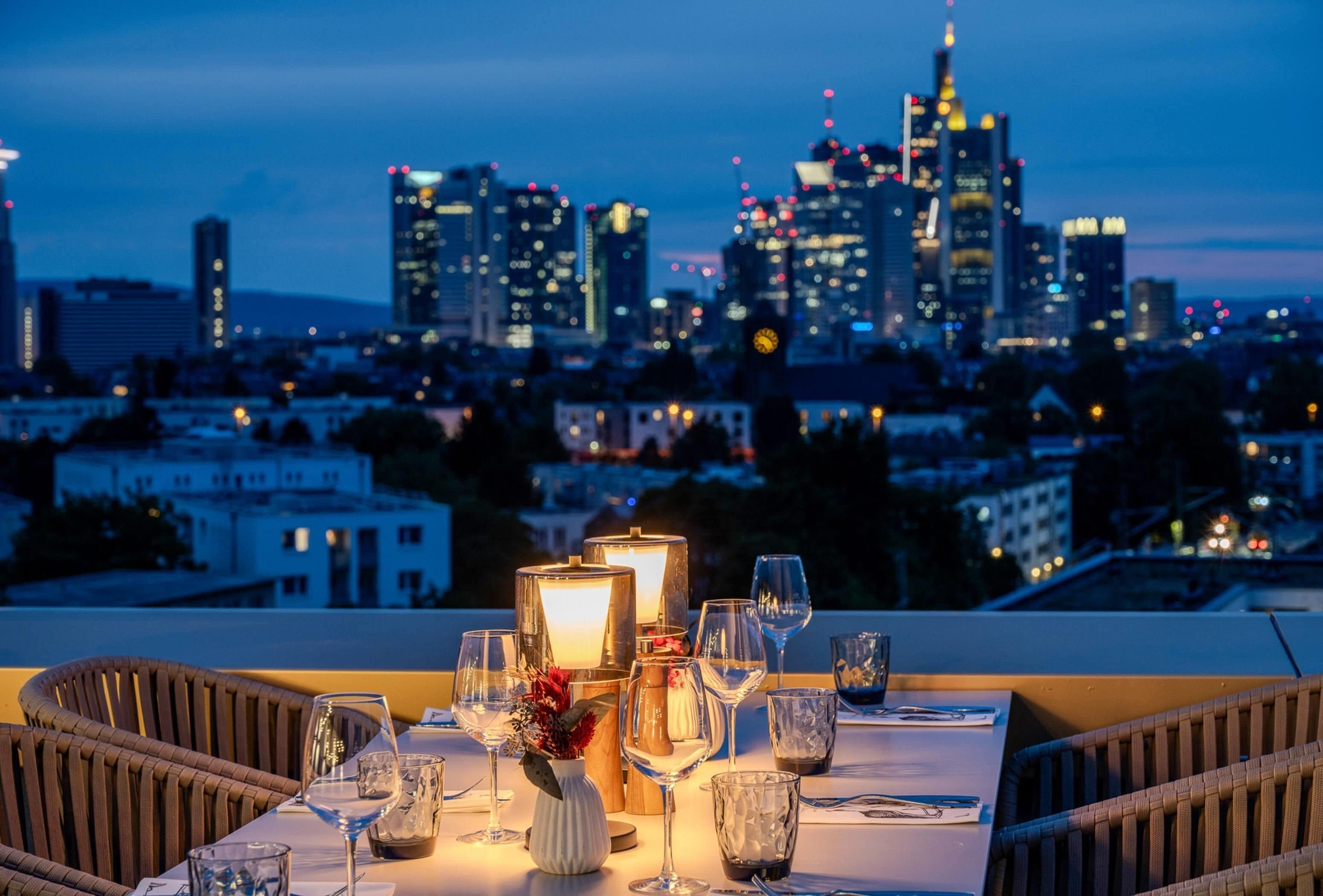 Een eettafel gedekt voor twee personen, met glazen en een kaars, met uitzicht op een stadszicht in de schemering.