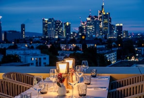 Un tavolo apparecchiato per due con bicchieri e candela, con vista sullo skyline cittadino al tramonto.