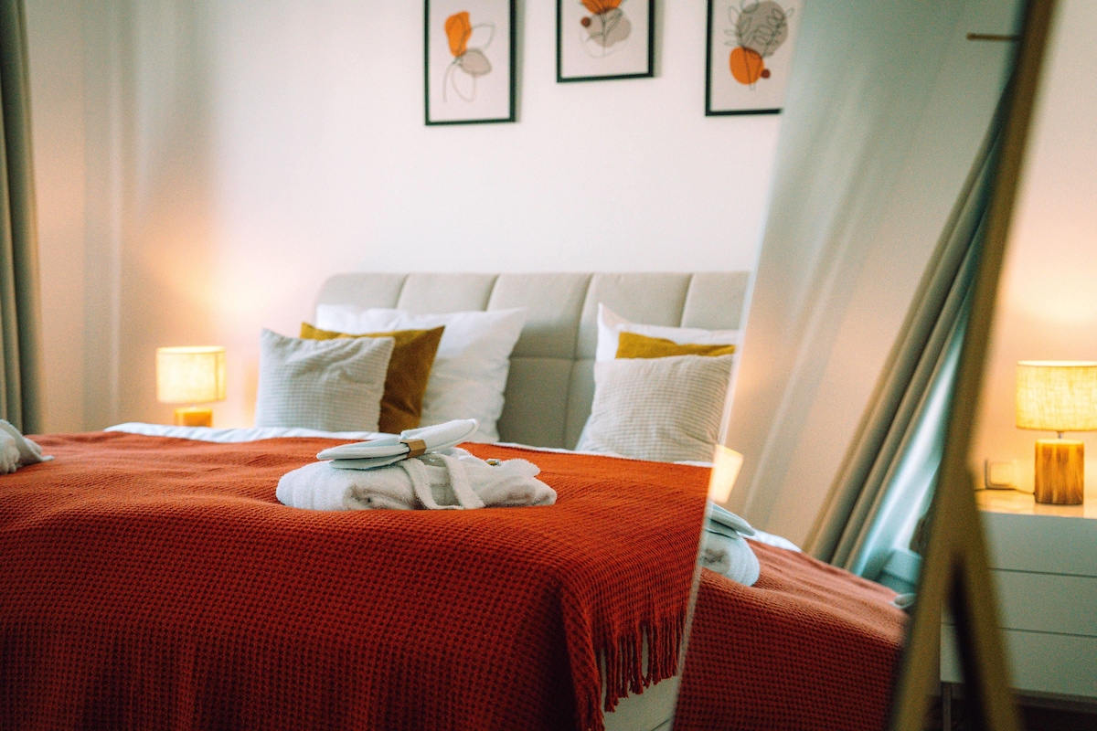 A neatly made bed with a red blanket, white pillows, and a white towel. There are framed pictures on the wall above the bed.