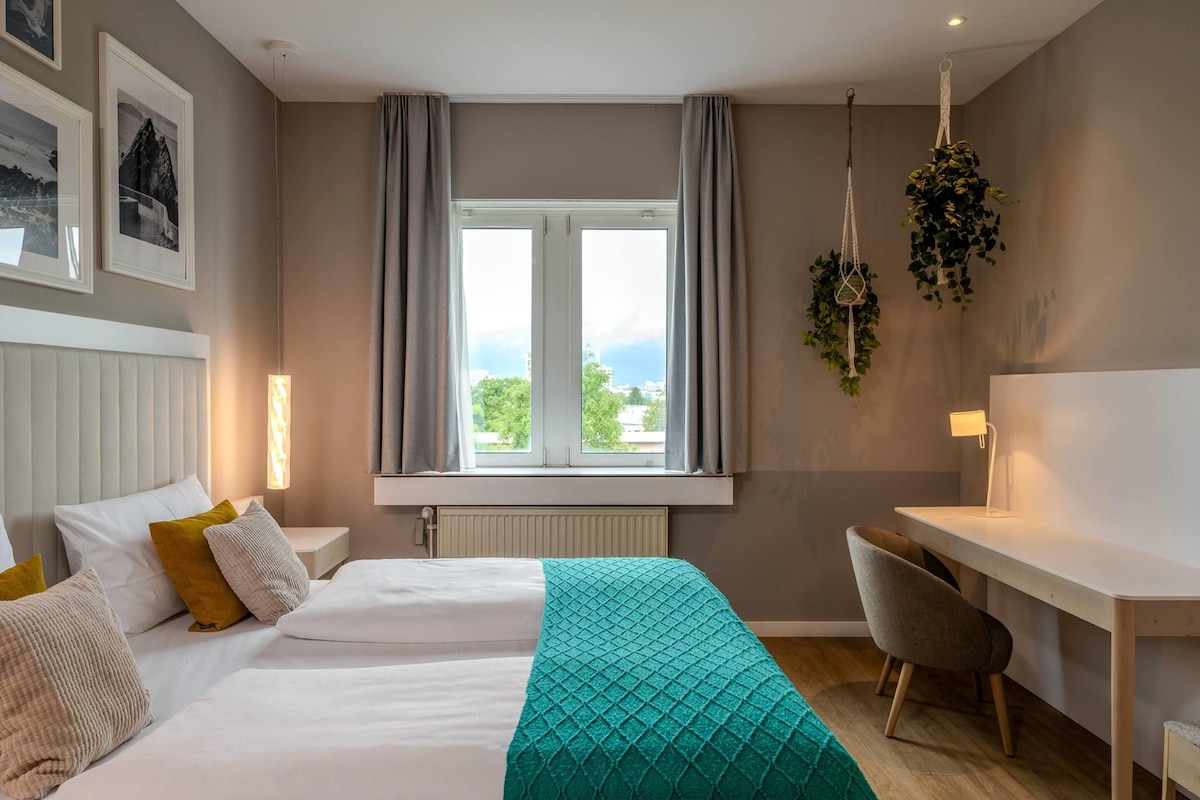 A hotel room with a bed, bedside table, desk, chair, and a window with curtains.