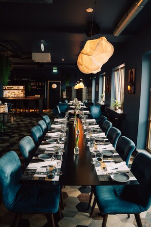 Ein Restaurant mit einem langen Tisch, der für eine Mahlzeit gedeckt ist, blauen Polsterstühlen und Pendelleuchten.