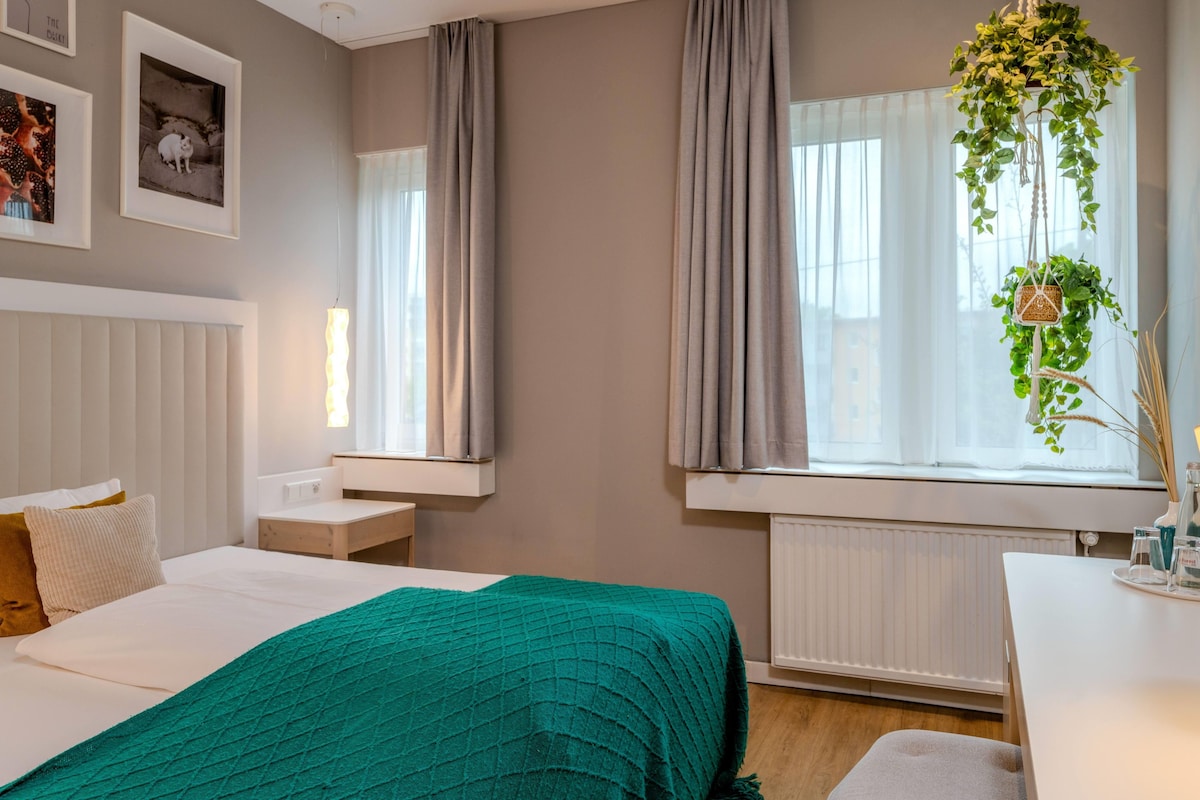 A hotel room with a bed, bedside table, a window with curtains, a radiator, and a wall-mounted plant.
