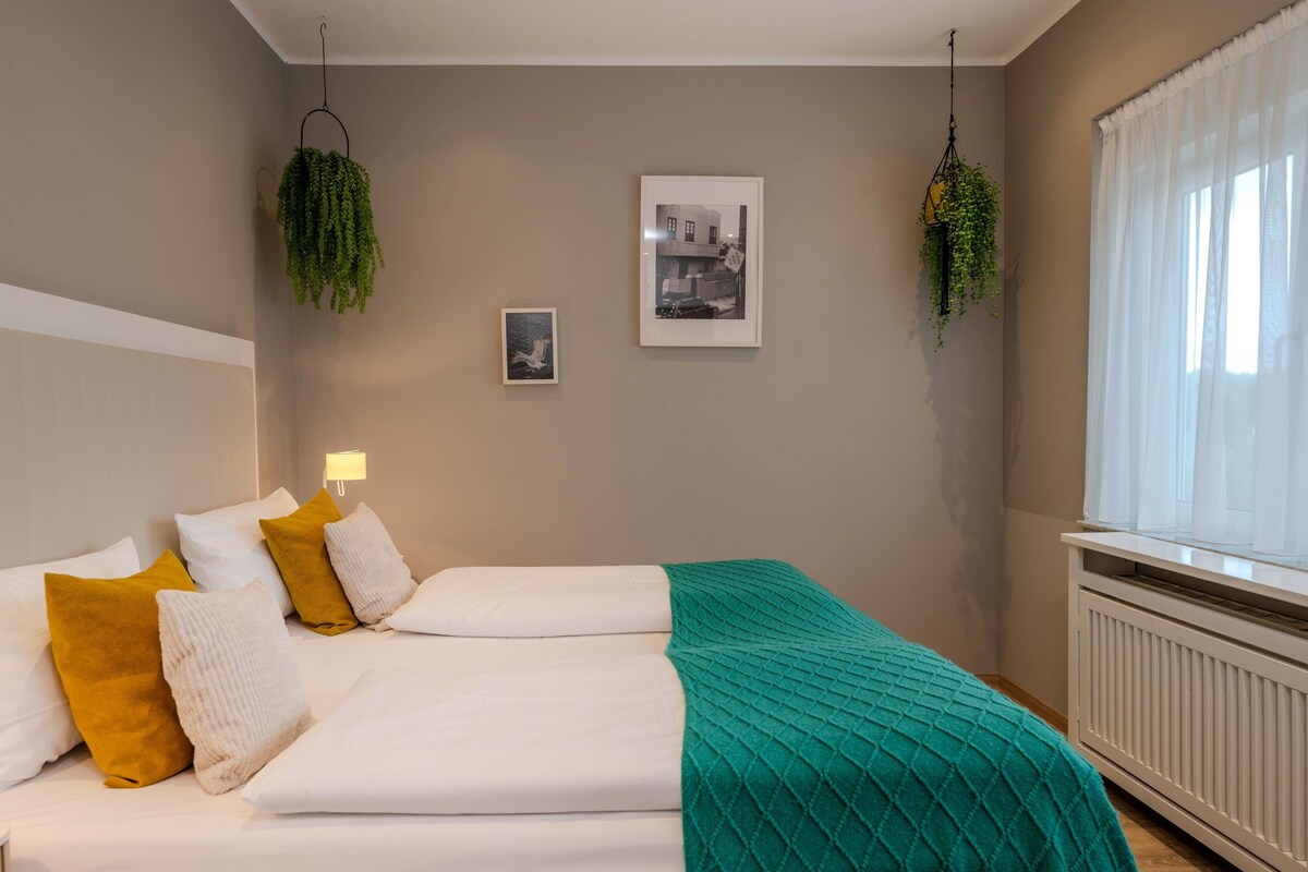 A neatly made bed with white and mustard pillows, a teal blanket, and a framed photograph on the wall.