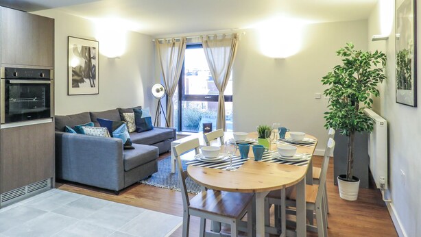 A modern living room with a sofa, dining table, and kitchen area including an oven. - Tudors eSuites Birmingham Apartments with Parking (Birmingham)