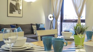 A modern living room with a sofa, a dining table with blue cups and plates, a floor lamp, and a window with curtains. - Tudors eSuites Birmingham Apartments with Parking (Birmingham)
