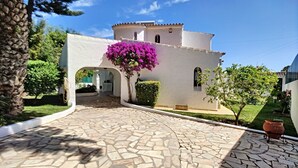 Un edificio blanco con entrada empedrada de piedra, un pequeño jardín y un árbol floreciente de color morado.