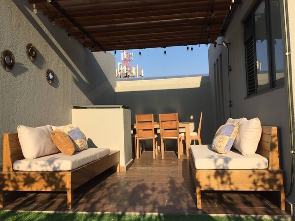 A patio area with two wooden sofas and a table, a pergola overhead, and a view of a building with a crane in the background.