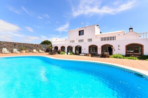 Un espace aménagé au bord d’une piscine à l’eau d’un bleu limpide, un bâtiment blanc orné d’arcades et un mur en pierre.