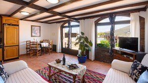A living room with a sofa, a dining table with chairs, a TV, and a large window with curtains. - Clara Suite 1: Large Pool, Spectacular Views, Terrace, Garden & Wi-Fi (Lanzarote)