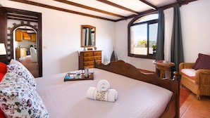 A bedroom with a large bed, a wooden headboard, a dresser, a mirror, a window with curtains, and a view of the outside. - Clara Suite 1: Large Pool, Spectacular Views, Terrace, Garden & Wi-Fi (Lanzarote)