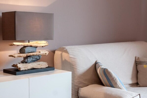 A modern living room with a minimalist design, featuring a white sofa, a stack of wooden logs on a black base, and a wall-mounted lamp.
