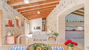 A dining area with a wooden table, chairs, and a bowl of fruit. There is a kitchen visible in the background. - Stone-villa in a rural idyll with pool– Villa Mamici (Capdepera)