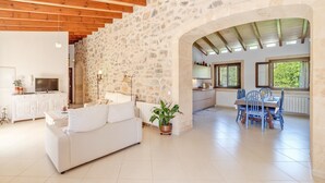 A spacious living area with a stone wall, wooden beams, a white sofa, a dining table with chairs, and a kitchen area. - Stone-villa in a rural idyll with pool– Villa Mamici (Capdepera)