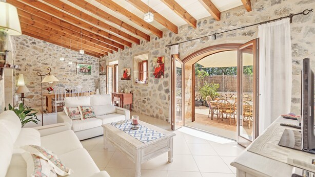 A living room with a stone wall, wooden beams, a white sofa, a coffee table, and a TV. - Stone-villa in a rural idyll with pool– Villa Mamici (Capdepera)