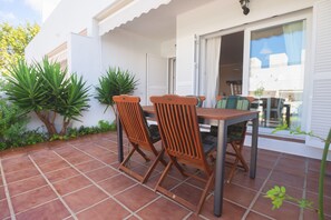 Une terrasse extérieure avec une table et des chaises, entourée de plantes et bordée d’un bâtiment blanc.