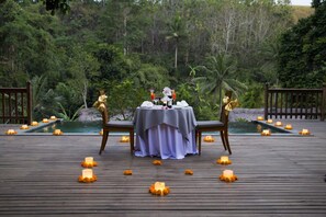 Une terrasse en bois avec une table dressée pour deux personnes, entourée de bougies et de fleurs, donnant sur une piscine et une végétation luxuriante.