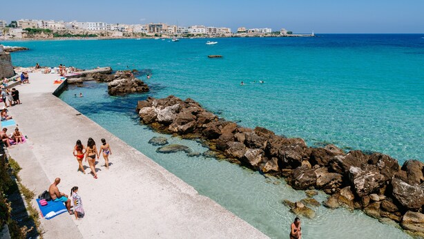 Una playa costera con agua cristalina, costa rocosa y edificios al fondo.