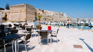 Terraza al aire libre con mesas y sillas, un edificio de piedra y un puerto deportivo con barcos.