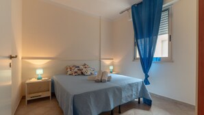 Un dormitorio con una cama, dos mesitas de noche, una ventana con cortinas y una puerta.