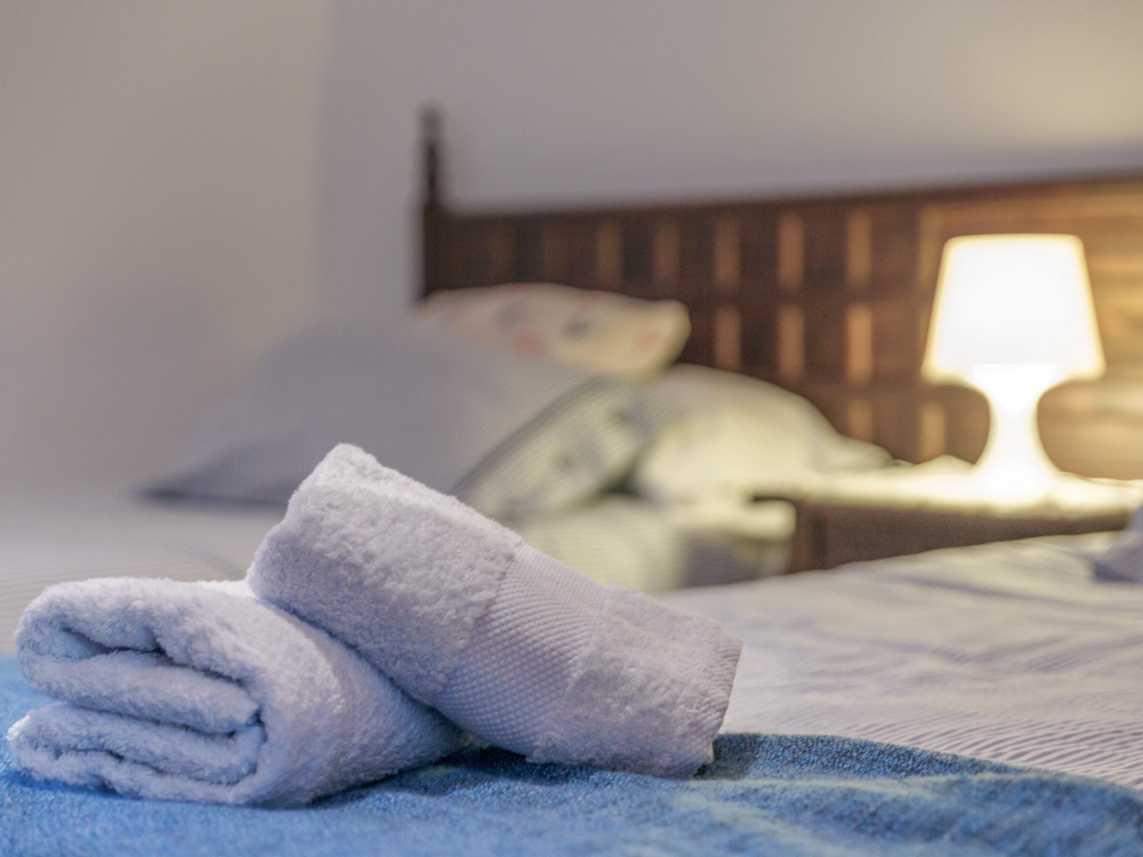 Una cama bien hecha con un edredón azul, una toalla blanca enrollada y una lámpara de noche.