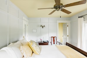 A bedroom with a bed, a wooden chair, a nightstand, a ceiling fan, and a door leading to another room. - Inn at Water's Edge (Plymouth)