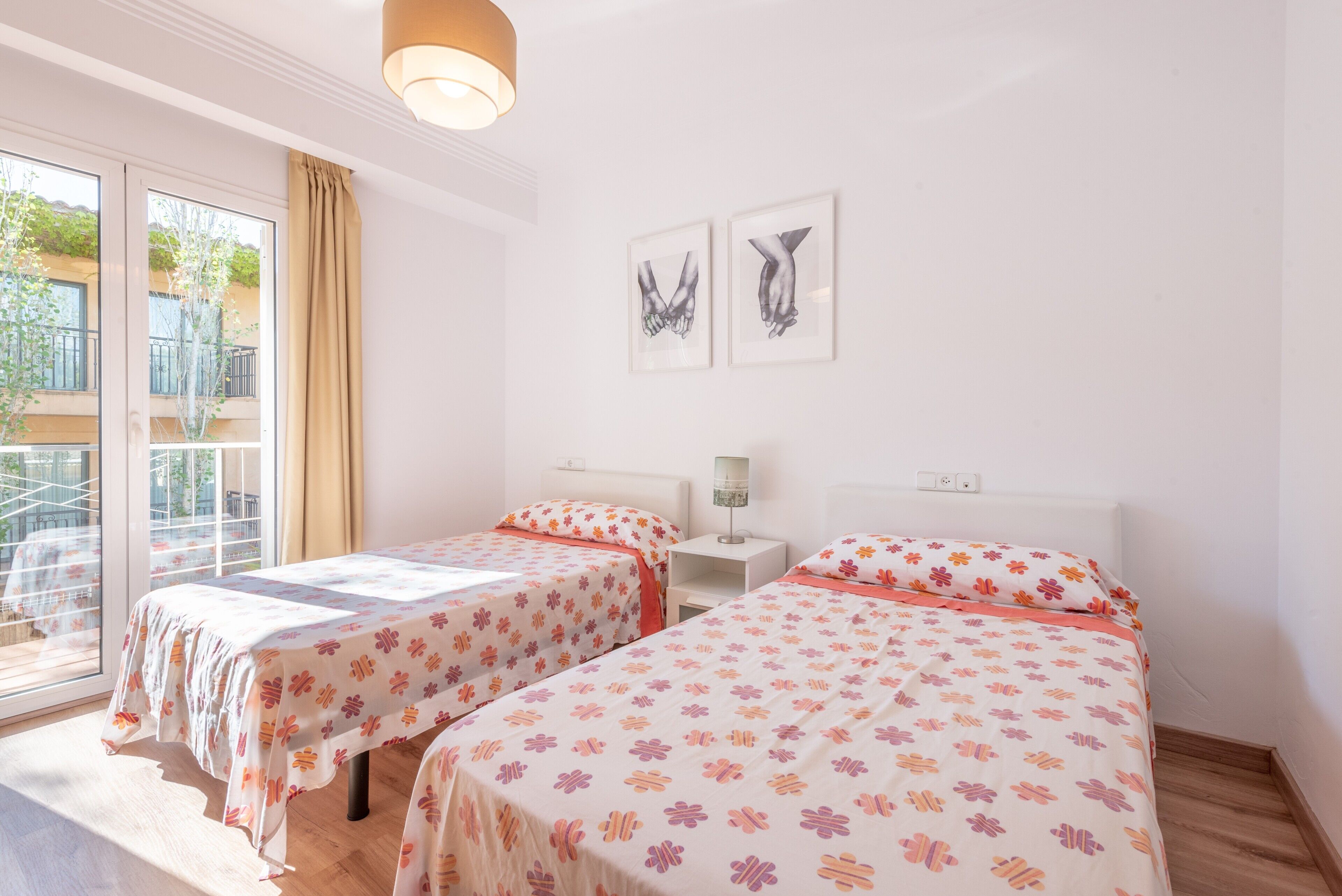 Ein Zimmer mit zwei Betten, einem Fenster mit Vorhängen, einem kleinen Tisch mit einer Lampe und zwei Wandbildern.