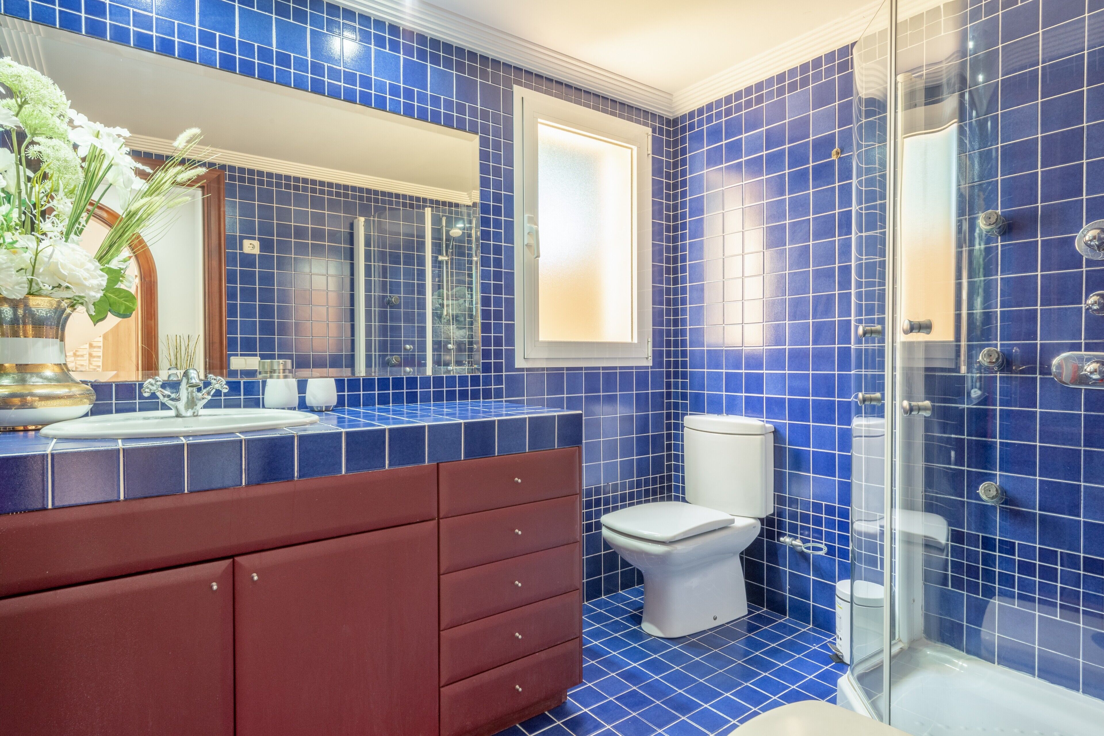 Een badkamer met blauwe tegels, een wit toilet, een glazen douchecabine en een houten wastafelmeubel met spiegel.