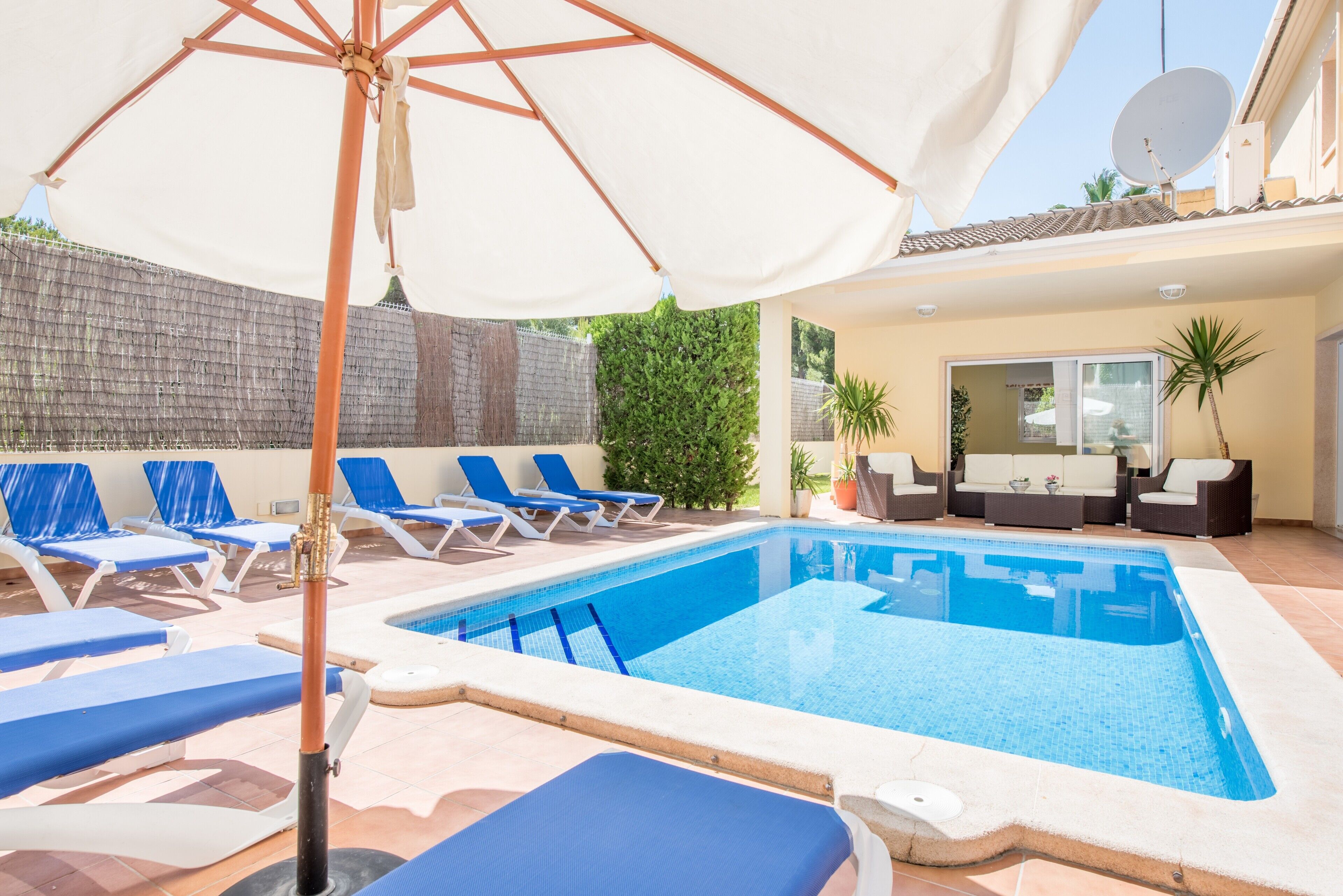 A swimming pool with blue lounge chairs and a white umbrella, surrounded by a tiled patio and a brick wall.