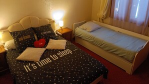 A bedroom with a bed, a smaller bed, a nightstand, a lamp, a wall art of a sunset, and a window with curtains.