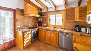 A kitchen with wooden cabinets, a built-in oven, a dishwasher, and a window with patterned curtains. - Rural villa with pool – Villa Tabo (Selva)