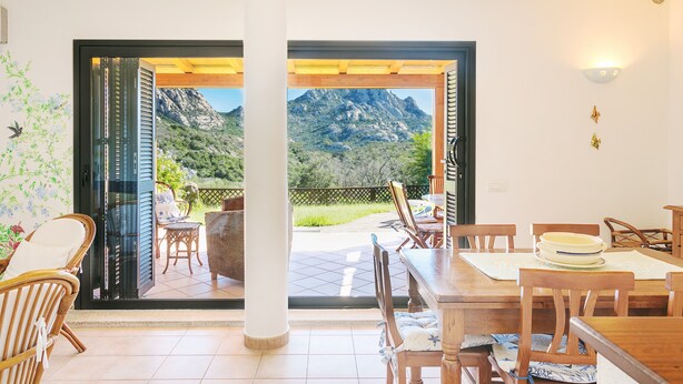 Uma sala de jantar com vista para montanhas através de portas abertas, mobiliário de madeira e chão em mosaico.