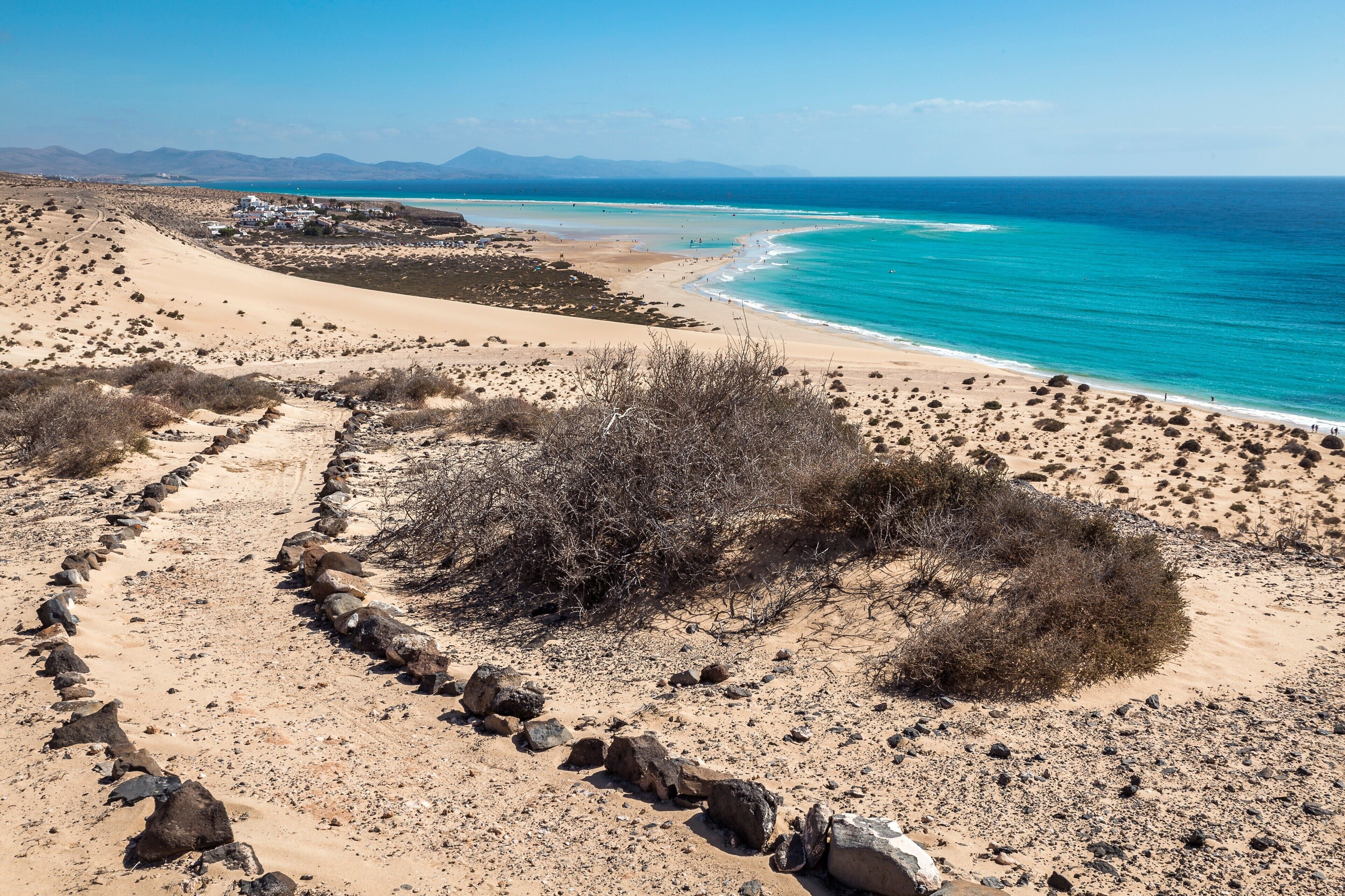 Een kustlandschap met een zandstrand, een heldere blauwe zee en een bergachtige achtergrond.