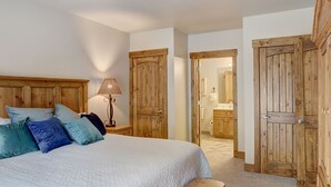 Quarto com cama, almofadas, abajur e portas de madeira que dão para um banheiro.