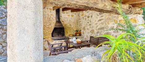 Un espacio al aire libre con paredes de piedra, una estufa de leña, un pilar de piedra y mobiliario de madera.