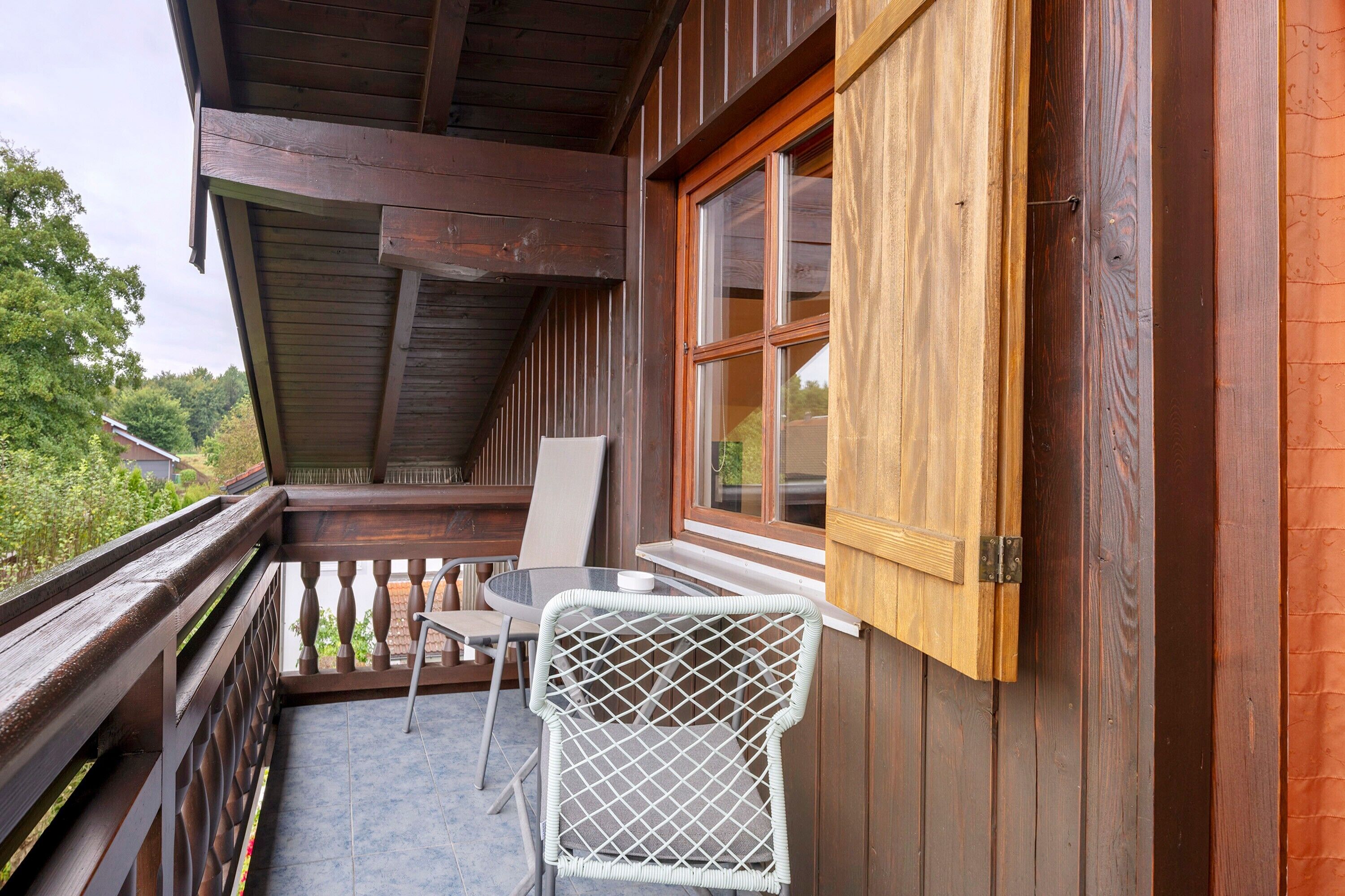 Un balcón de madera con una mesa y sillas, una ventana con contraventanas y vistas al verdor.