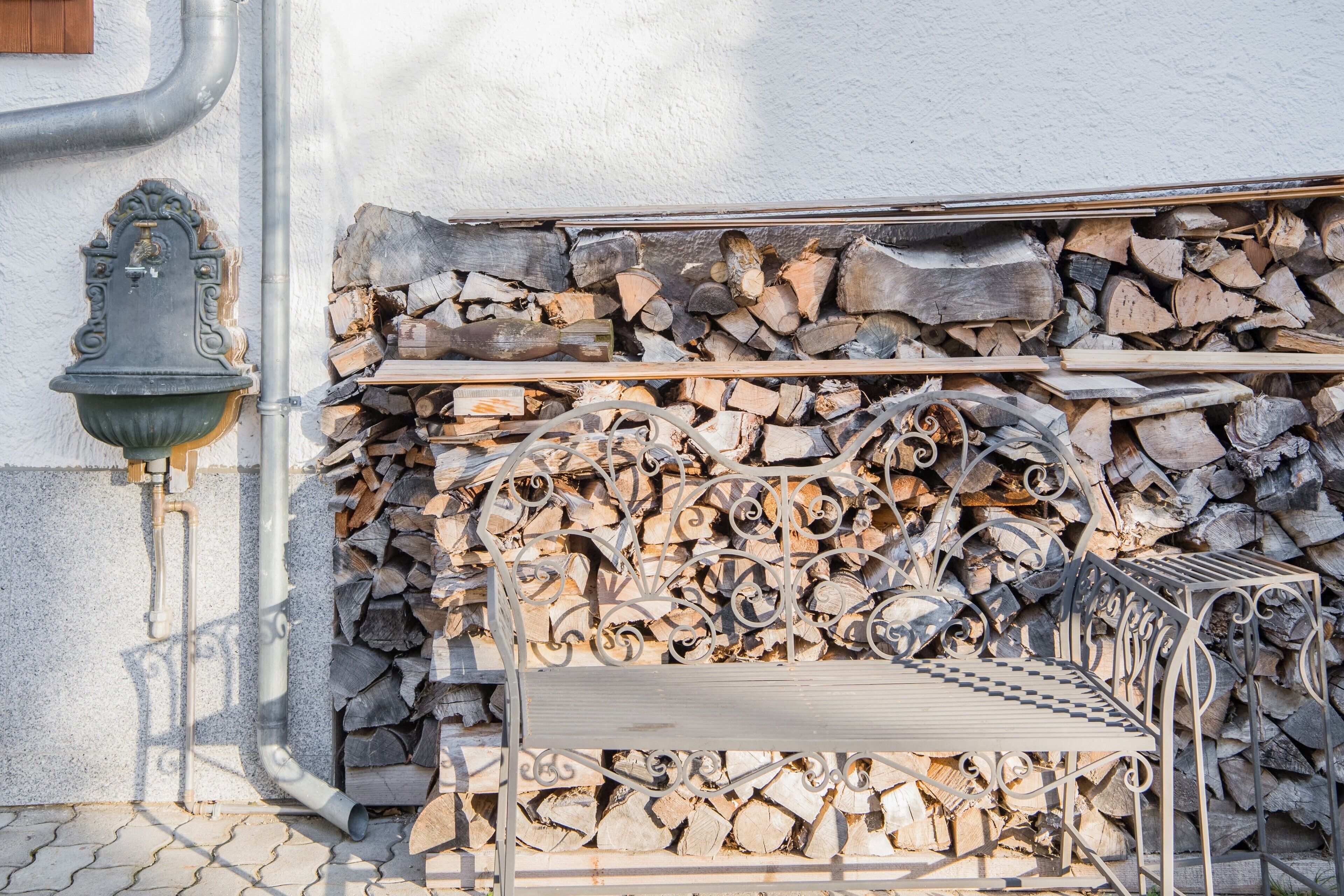 A metal bench with a checkered pattern, a metal railing, and a stack of firewood against a wall.