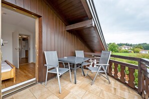 An outdoor balcony with a wooden railing, a table with chairs, and a view of a green landscape.