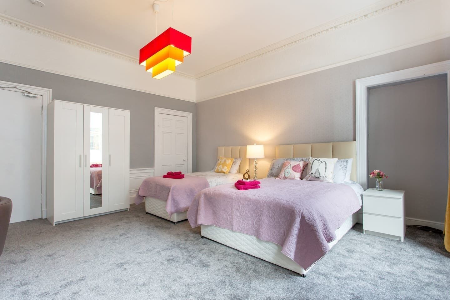 A bedroom with two beds, a wardrobe, a bedside table with a lamp, and a modern pendant light.