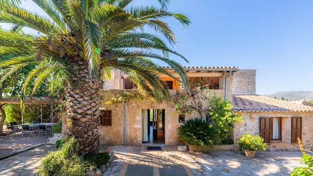 A two-story stone house with a stone staircase, a palm tree, and a patio area.