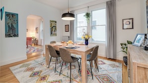 A dining area with a wooden table, four chairs, a hanging light fixture, and a window with curtains.