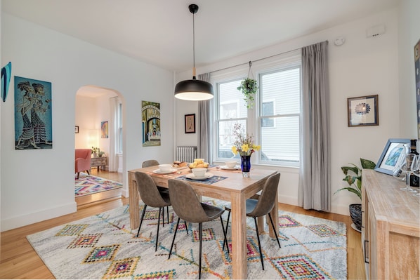 A dining area with a wooden table, four chairs, a hanging light fixture, and a window with curtains.