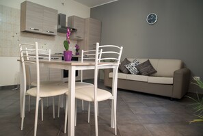 A modern kitchen with white cabinets, a dining table with four chairs, and a beige sofa in the living area. - Nick & Santo’s Holiday Homes C1 (Giardini Naxos)