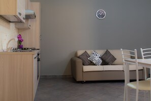 A kitchenette with a stove, a purple orchid, and a beige sofa with patterned pillows. - Nick & Santo’s Holiday Homes C1 (Giardini Naxos)