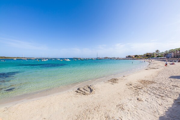 A beach with clear turquoise water, a sandy shore, and a few boats in the distance.